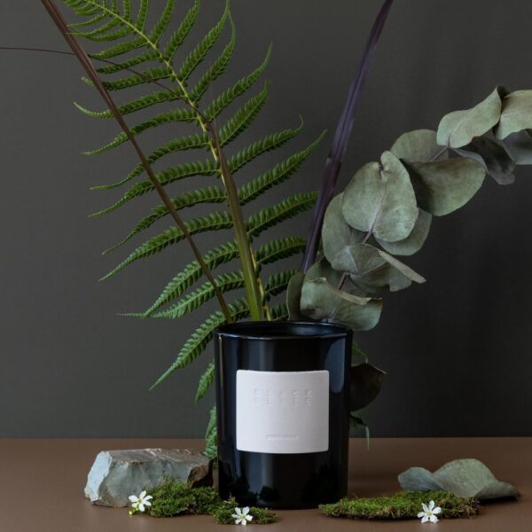 A cylinder-shaped jar of candle placed in front of a blooming flower and its leaves.