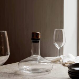 Natural light, perspective view of a glass wine breather next to wine glasses on top of a table.