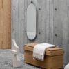 Dowel large oval mirror on a concrete wall with a beige sculpture on the floor and a blanket on the oak bench