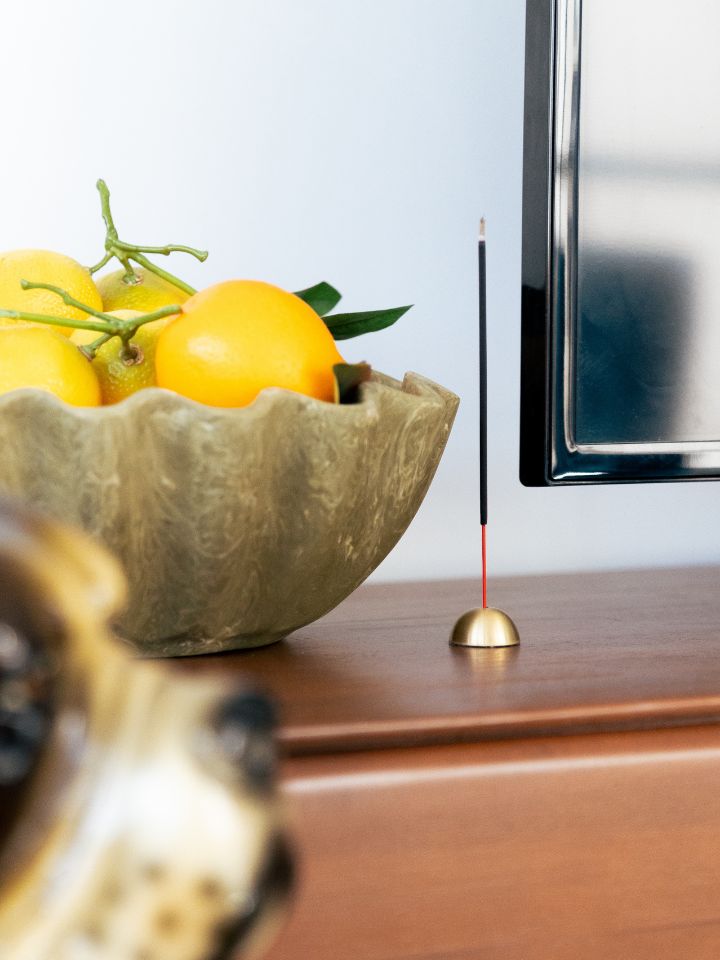 A Behr & Co brass incense holder with a stick of burning incense, next to a fruit bowl.