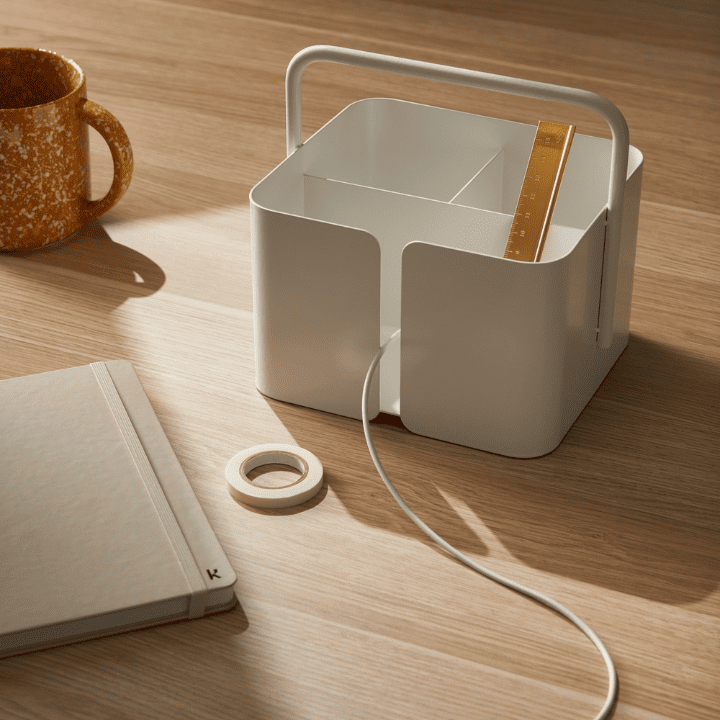 This photo illustrates the 2023 design trend of Multifunctional Spaces and features the Designstuff Carry-All Caddy. The Caddy is white and holds a brass ruler, and a cable is protruding from a small thin gap. It's set on a timber work desk, and has been placed near a notebook, some tape, and a speckled coffee mug.