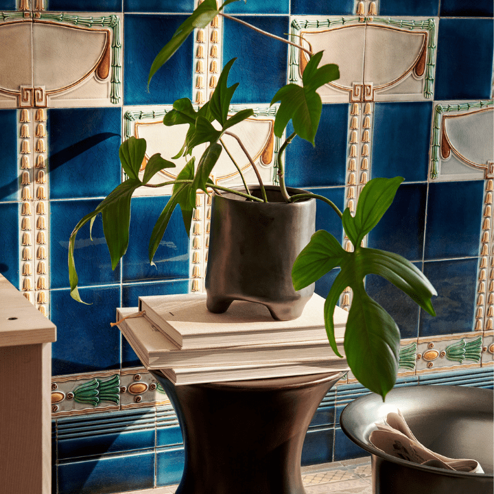 This photo illustrates the 2023 design trend of Biophilic Design, and shows a photo of ferm LIVING's Esca pot with a big leafy green plant insite it. It's sat atop a pile of books and a brown timber stool. Behind it is a ornately tiled wool featuring a deep blue tiles.