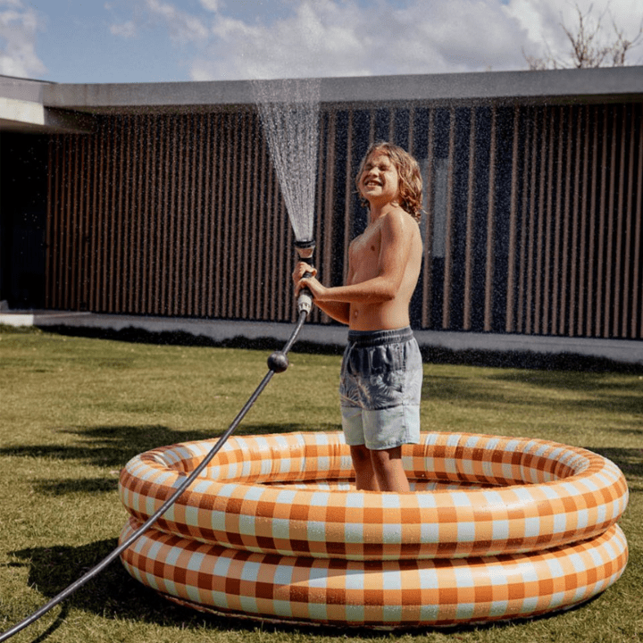 The photo is of a kid standing in a Pool Buoy pool, spraying water into the air through a hose. The inflatable pool is in a sky blue and orange gingham pattern.