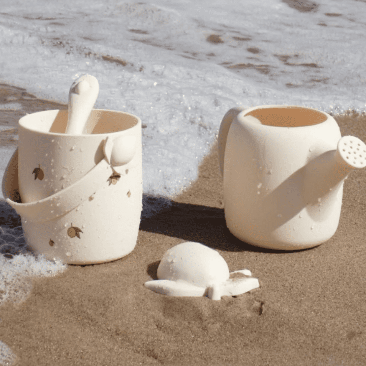 The photo is of the Konges Slojd silicone beach set in a light yellow colour. The toys include a bucket, watering can, and mould. They're placed on the sand, with a foamy wave behind.