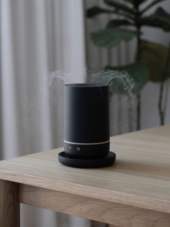 A photo of the 360 Diffuser, designed by PerCapita for In Essence. The shot is lifestyle based and shows the black tubular unit on top of a simple timber table. Smokey white vapour can be seen coming out in all directions from the design's top opening.