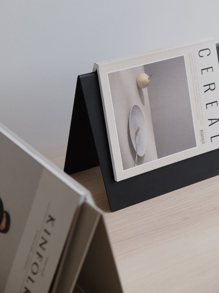 The photo is a product shot of the Bookmark and Magazine rest designed by PerCapita. The units are triangular shaped and stand with the pointy end facing up. On top of the magazine stands, one black and one beige, are open magazines, one is Cereal and the other is Kinfolk.