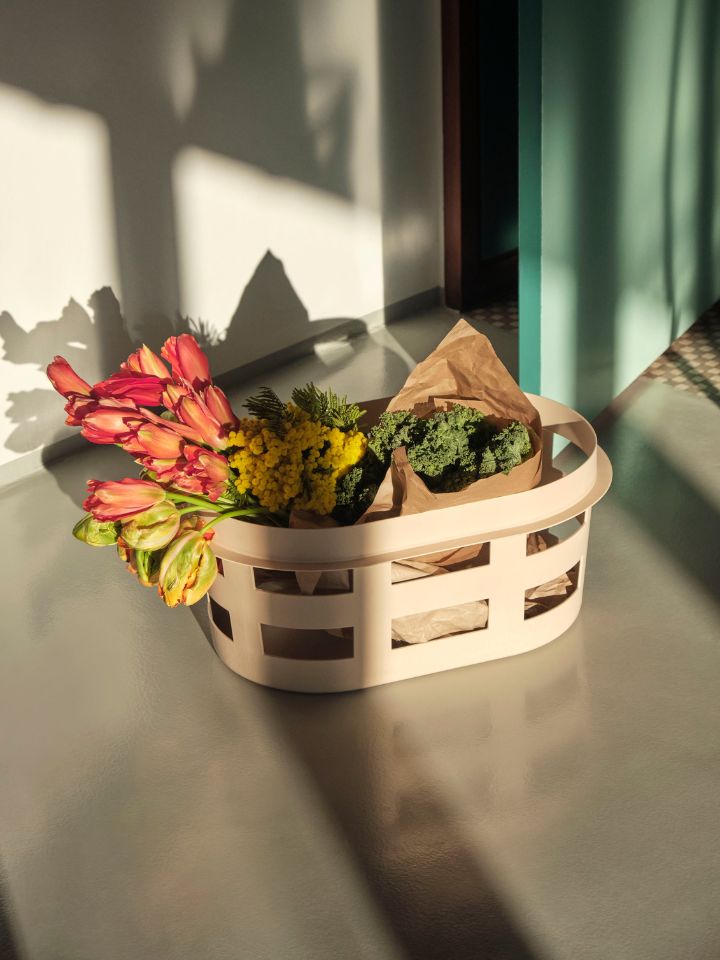 A photo of HAY's laundry basket in a soft beige yellow colour. The basket is on top of a polished concrete floor. Inside the basket are bright flowers and greenery, wrapped in brown paper. The lighting here is moody, with plenty of shadow play cast by the basket and florals.