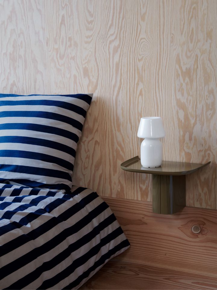 This is a photo of a bedroom, featuring HAY's Korpus shelf in an army green colour. The glossy shelf is attached to a grained timber wall, holds a glossy white bedside lamp, and is next to a thickly striped blue and white bedspread.