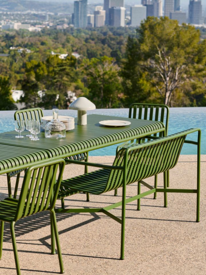 An outdoor shot of the HAY outdoor Pallisade range, including a table, chairs, and bench, in a muted army green tone. The setting is alongside an infinity pool, overlooking plenty of trees and a city skyline visible in the distance. On top of the setting are a number of glasses, plates and carafes, arranged neatly. There is also a glass Pao lamp in a soft grey on top of the table.