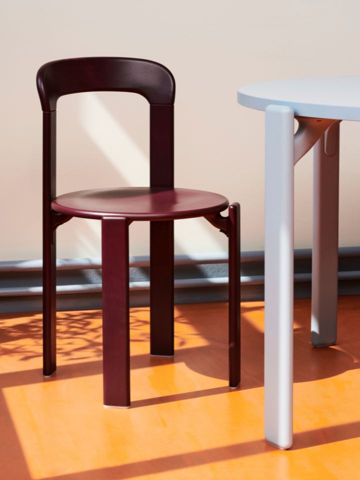 A photo of HAY's Rey Chair in a deep grape burgundy. It sits alongside a pale blue table, on top of a vibrant orange floor. The lighting is moody, and both the table and chair casts a dark shadow.