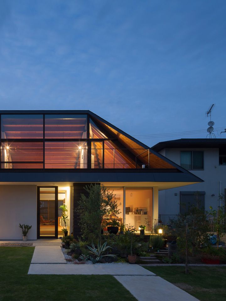 The photo is of the House With Large Hipped Roof, a project by Naoi. The shot has been taken at night time, which shows of the home's large hipped roof and its floor to ceiling windows, which are illuminated against the dark.