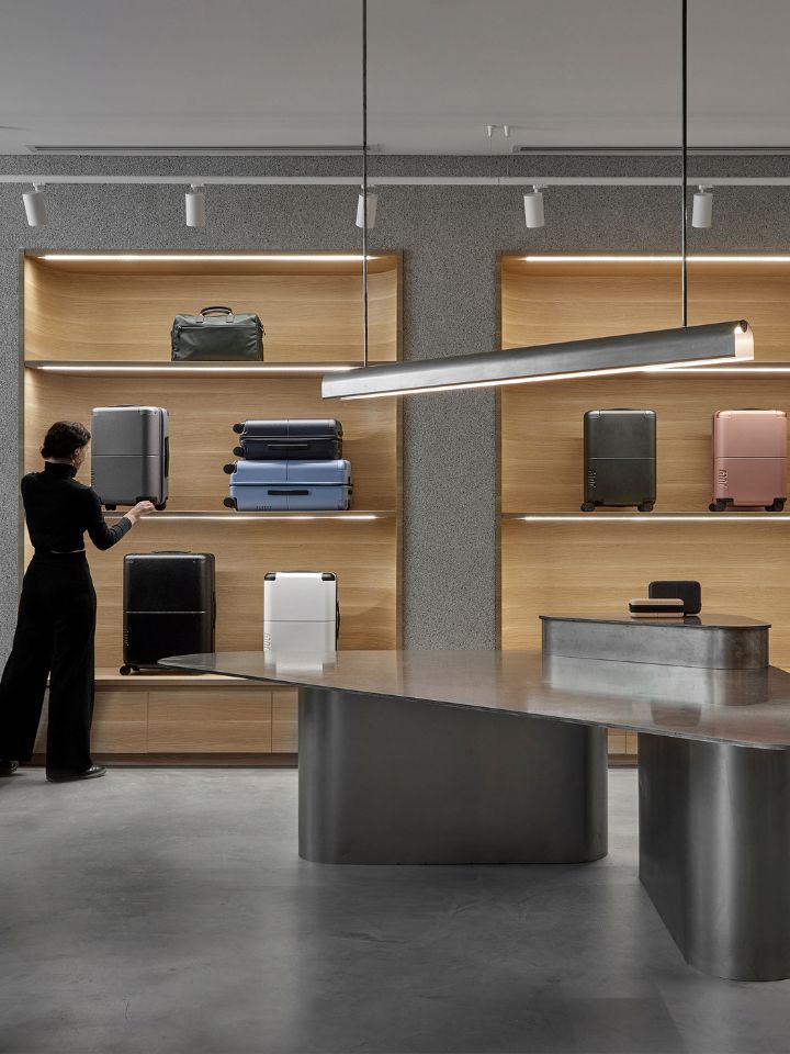 A photo of July's Flagship store, taken by Dave Kulesza. The interior of the store is clean and minimalist, similar to the product the brand sells. There are neatly stacked and displayed pieces of luggage, which are being attended to by a woman dressed in black. IN the centre of the store is a glossy metallic table with a large pendant suspended above.