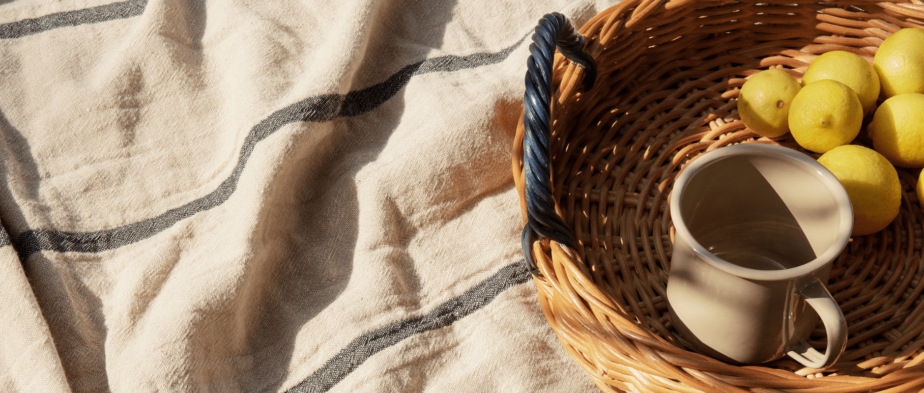 A photo of ferm LIVINGs Yard Picnic Blanket in a beige colour with a black stripe with a basket sitting on top with a mug and lemons inside the basket.
