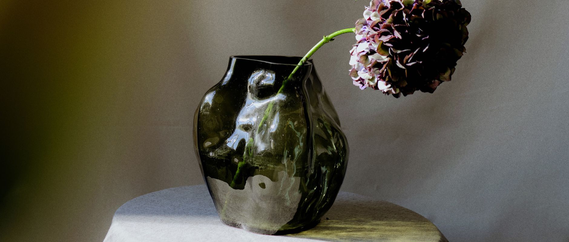 Banner for a blog post about how to achieve a timeless and elegant look in your home. The image features NEW WORK's glass Blaehr vases, which has a unique organic shape and is speckled with indents. The vase is sitting on a table with a white table cloth and has a dusty purple flower in it.