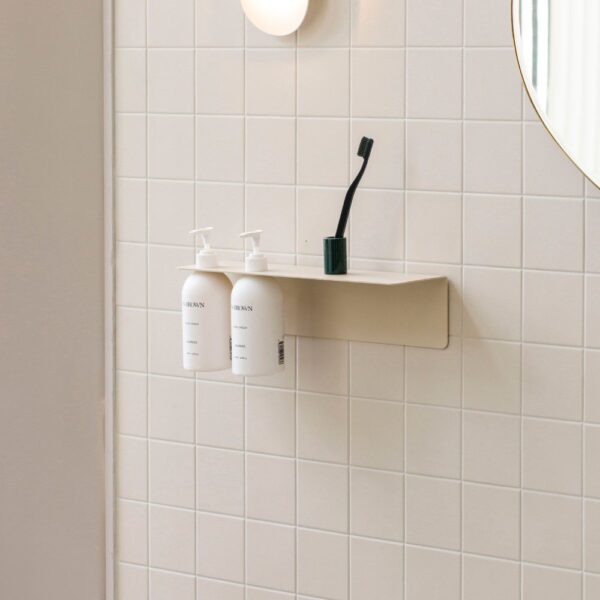 Natural light, perspective view of a white soap dispenser holder mounted on a wall with two bottles of soap attached and toiletries placed on its shelf.