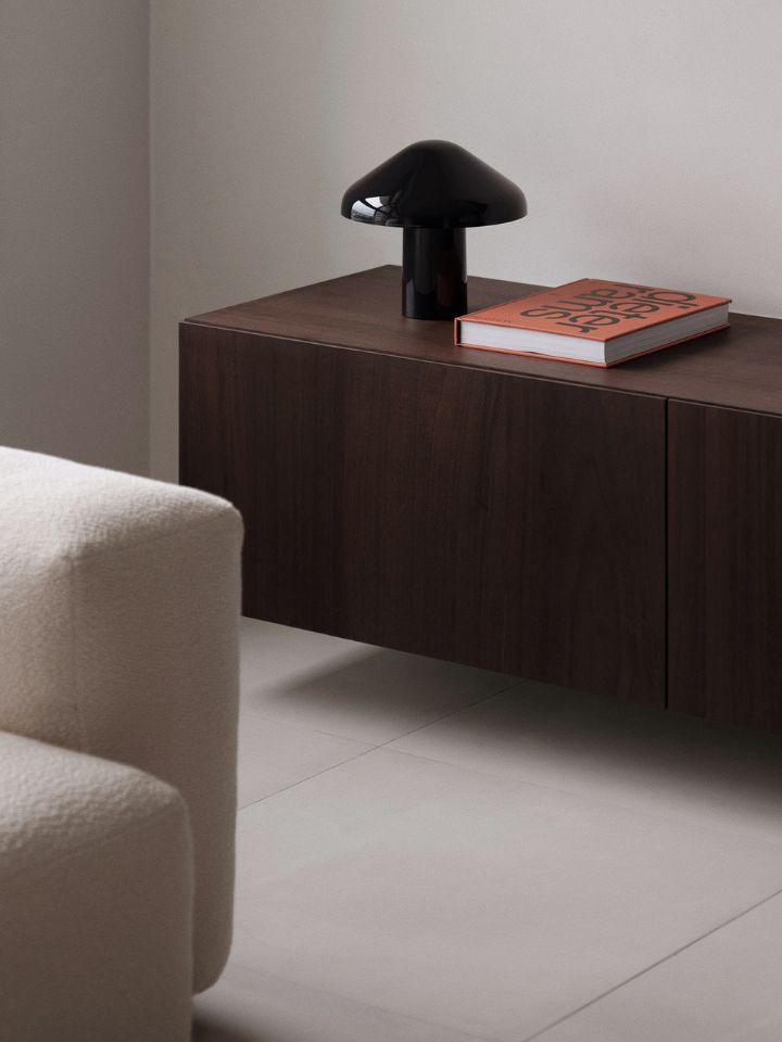 Photo is of an internal shot of the East Coast Residence in Singapore. This particular shot shows a floating dark timber cabinet, with a red book and black table lamp sitting on top. The unit is attached to an off-white wall, and there is a cream boucle sofa to the far left of the image, only just in focus.