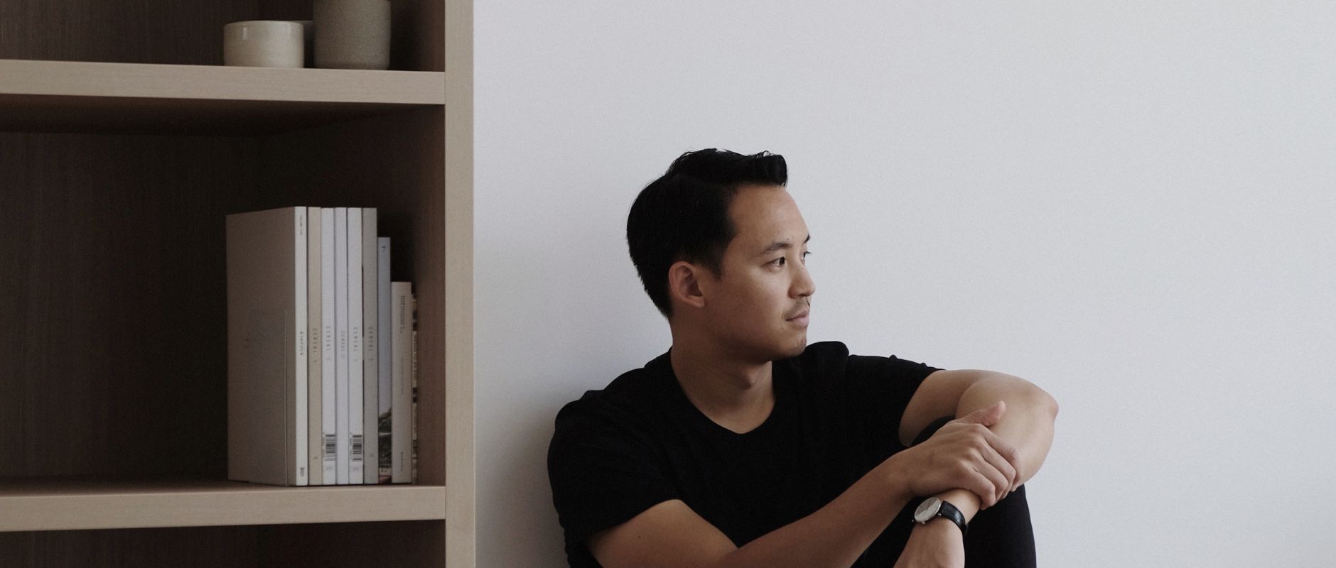 The photo is a profile shot of industrial designer, Jon Liow - the subject in this interview. He's wearing a black tee-shirt and is sitting casually on the ground with his arm resting on his knee. He's looking to the right hand side of the photo, and on his left is a bookshelf with a tidy stack of books on it.