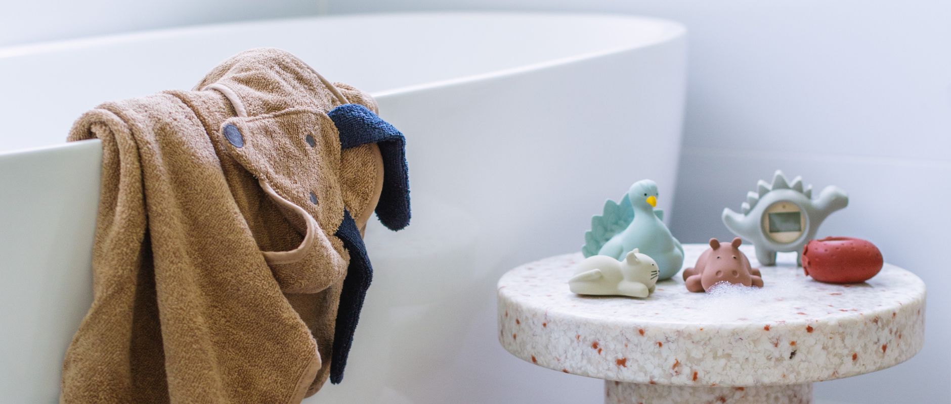 This image is of a cute bath set up for a young child or toddler. It shows a bath tub with a dog shaped hooded towel draped over the side (by LIEWOOD), a speckled side table made by NORMANN COPENHAGEN, and plenty of colourful bath toys and thermometers by LIEWOOD, NATRUBA and KONGES SLOJD. The bath toys are shaped as animals and include a cat, a peacock, and a hippo. There are plenty of soap suds everywhere.