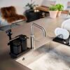 Natural light, isometric view of a stainless steel kitchen sink with a black-coloured tray of cleaning tools beside the sink's faucet.