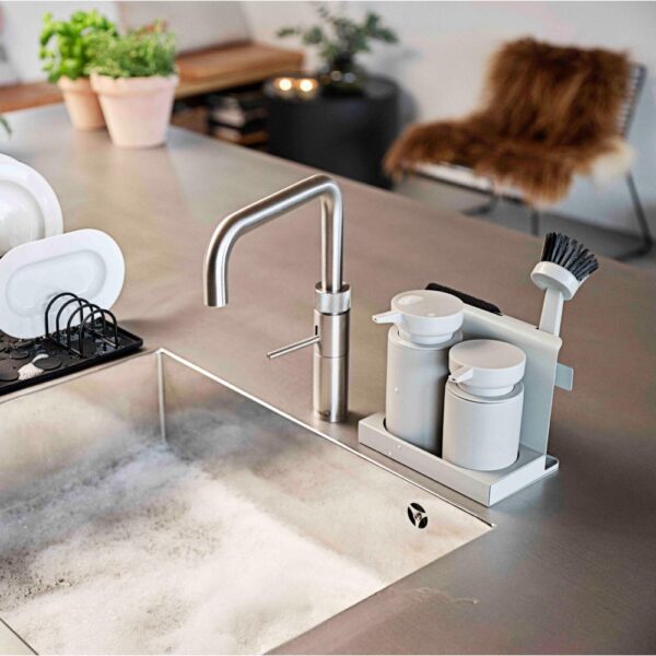 Natural light, isometric view of a stainless steel kitchen sink with a grey-coloured tray of cleaning tools beside the sink's faucet.