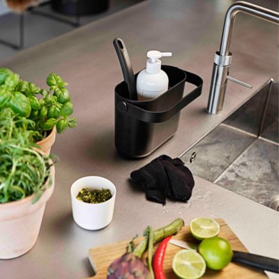 Natural light, isometric view of a stainless steel kitchen sink with a black-coloured tray of cleaning tools beside the sink's faucet and a chopping board with sliced fruits and vegetables.