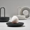 An egg perched on a black egg cup with other egg holders in the background