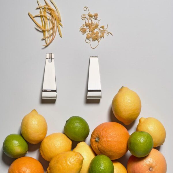 Grey background, studio image of two polished, silver-colored steel channel knives with a bunch of citrus fruits laying beside them.