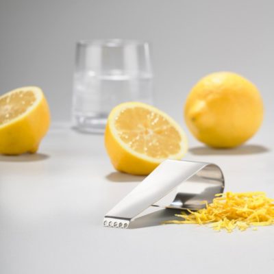 Perspective view of a julienne knife on the foreground with three sliced lemons and a glass of water in the background.