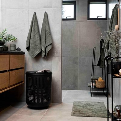 Natural light, perspective view of a black, cylindrical laundry basket on the floor in a concrete-walled bathroom with two towels hung above it.