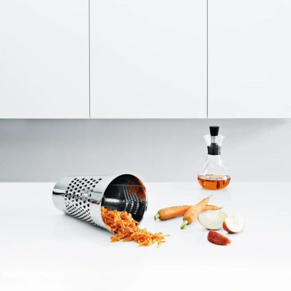 White background, studio lighting, perspective view of a stainless steel, canister-shaped grating bucket with shredded carrots spilling and a jar of salad dressing in a kitchen counter.