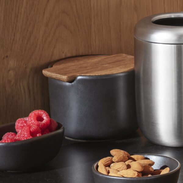 Natural light, perspective view of a black, canister-shaped steel salt cellar with a bowl of nuts in the foreground.