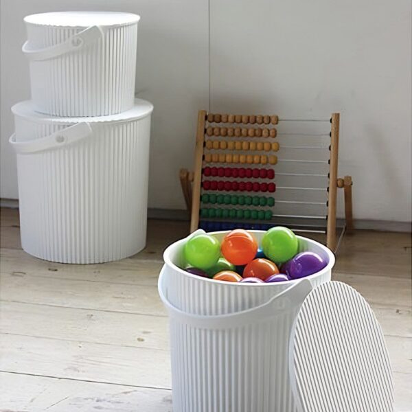 Natural light, perspective view of a white storage bin with multi-coloured balls inside of it and two white storage bins stacked in the background.