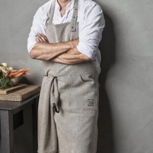 Studio lighting, unpainted grey concrete background and a person with an arms crossed pose, sporting a grey apron over a white shirt.