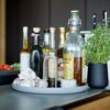 Natural lighting, perspective view of a kitchen counter with bottles of condiments and spices gathered inside a grey circular tray with a black flower pot with herbs, beside it.