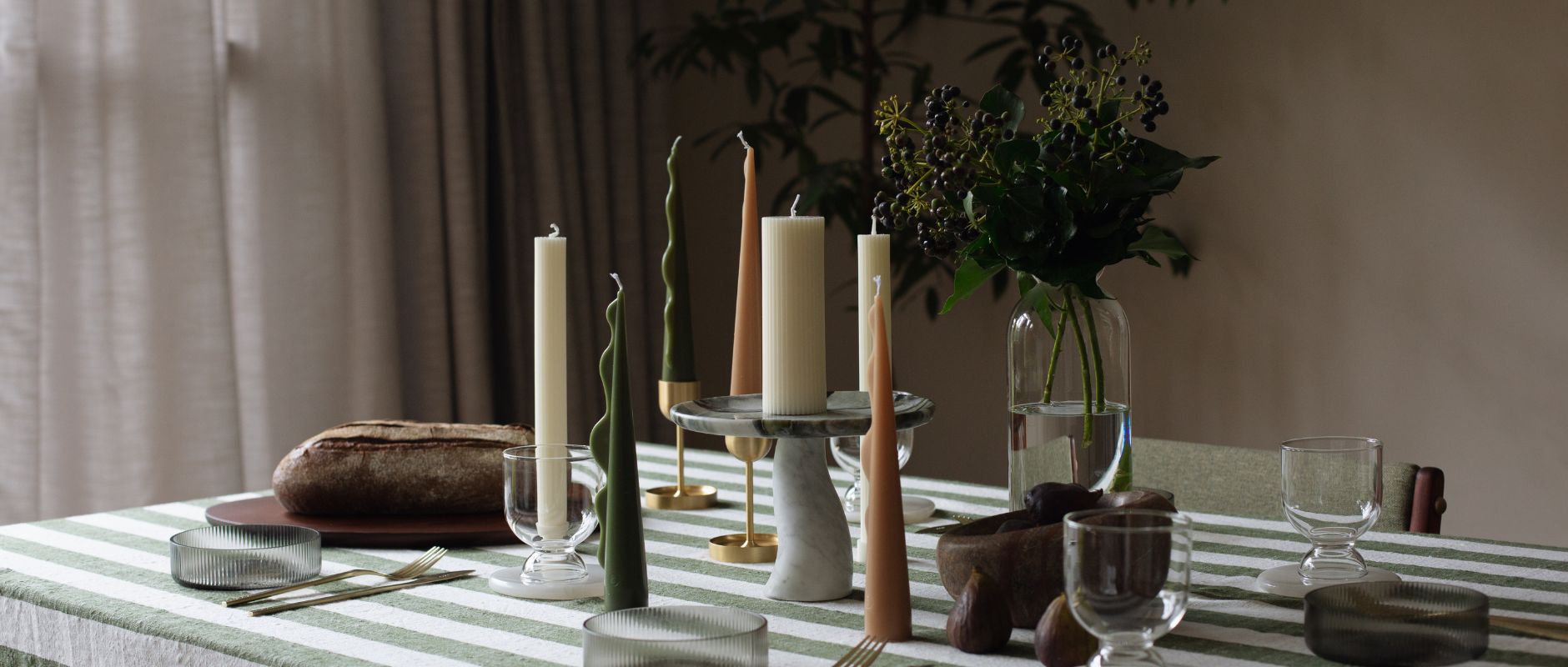A banner showing a beautiful table setting featuring candles and home decor by Black Blaze. The table has an Oyoy tablecloth in a green and white stripe, trays, bowls and candles by Black blaze, in neutral tones of olive, toffee, and off-white. There are also sculptural glasses scattered around the table by ferm LIVING. The mood of the table setting is soft and ambient.