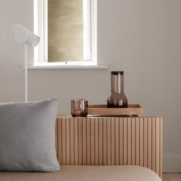 A wooden tray with a cup and glass carafe placed on top of a table next to a bed and pillow.