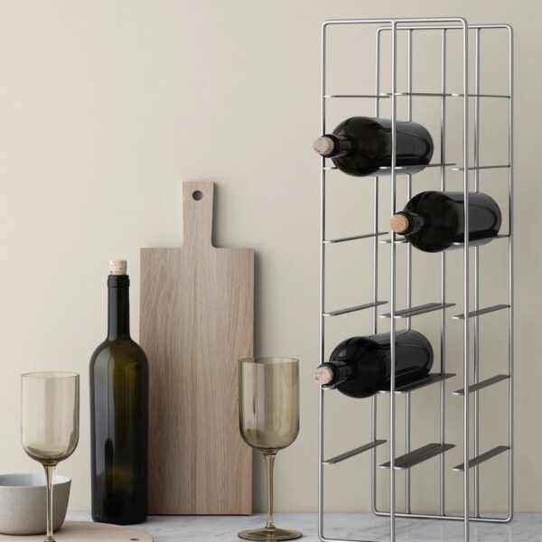 Three bottles of wine stored and stacked in a grid-type container placed next to an upright wine bottle, cutting board and wine glasses.