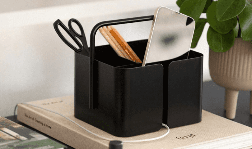 The Designstuff desk organisation caddy holding an assortment of stationery and a phone charging. The charging cable protrudes from the caddy's cable slit.