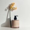 Studio lighting, perspective view of a hand moisturiser bottle placed next to a flower stem leaning back on a wall.