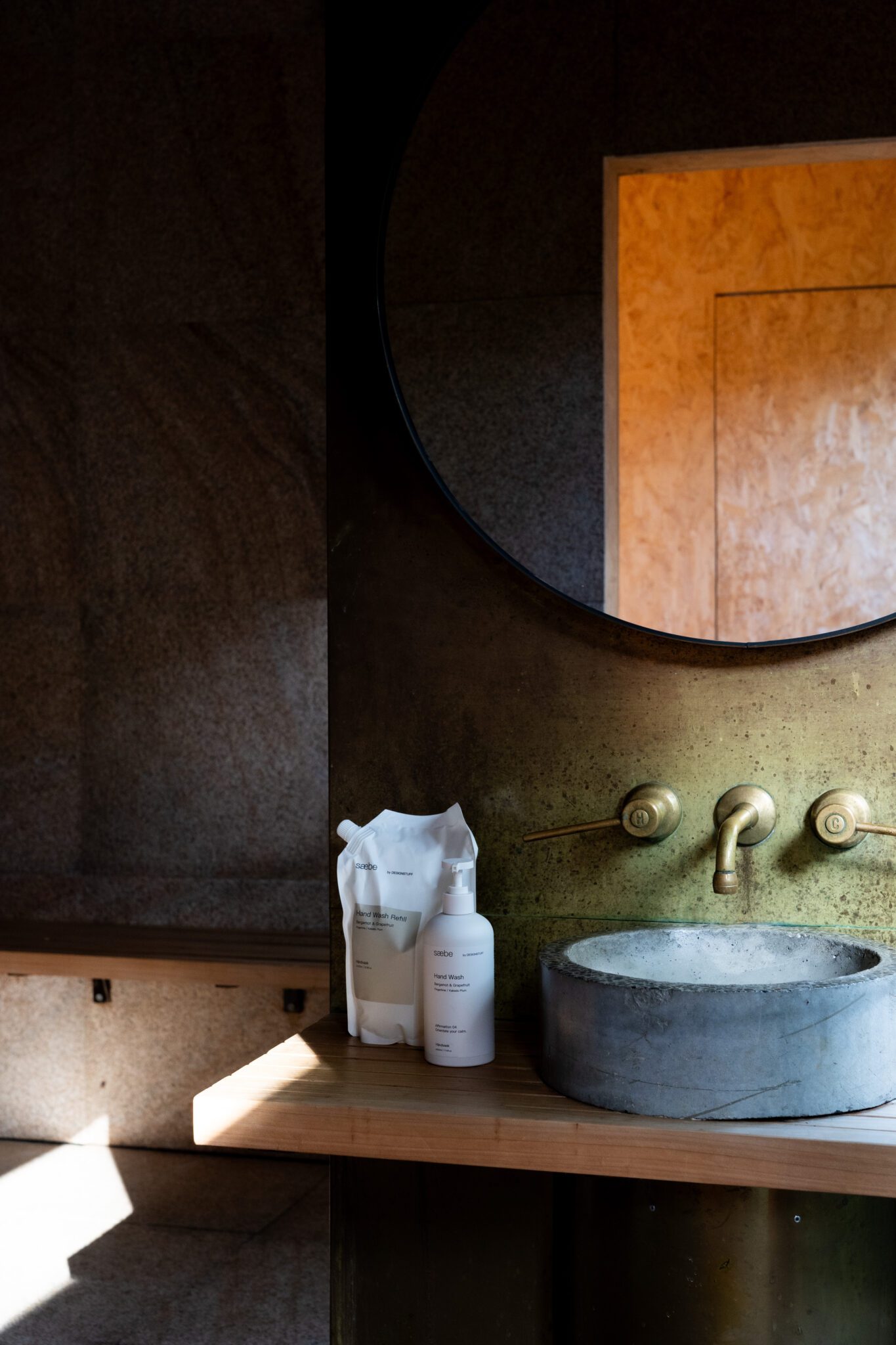 saebe by Designstuff hand wash and saebe by Designstuff hand wash refill on a bench in a spa-esque bathroom next to a stone sink with brass details and large round mirror.
