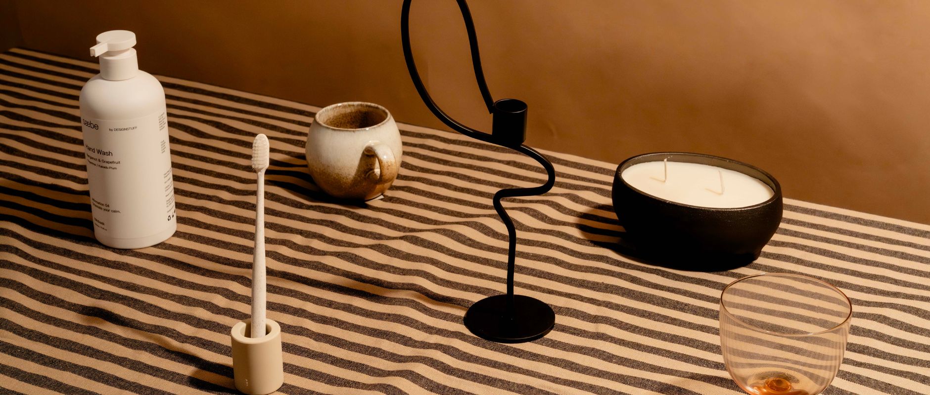 A beige-themed gift guide displayed on a striped brown and black tablecloth. Featured items include a hand wash bottle, toothbrush in a holder, sculptural candle holder, candle in a pot, and a blush-tinted glass.